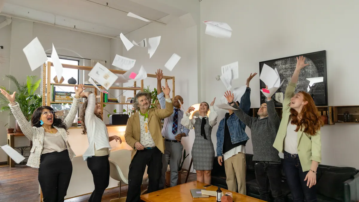 Un groupe diversifié de collègues de bureau célèbre en lançant des papiers en l'air ; des hommes et des femmes rient.