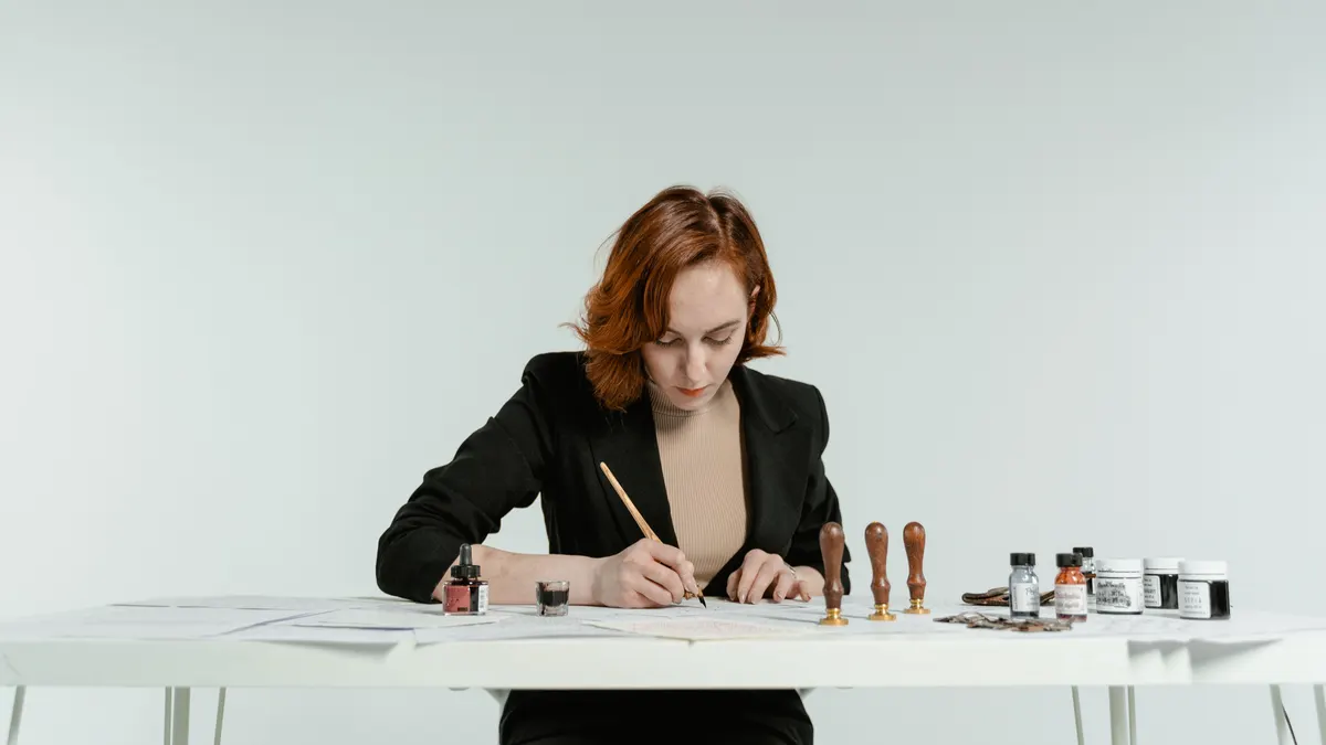 Femme caucasienne en blazer noir tenant une plume trempée pour écrire à une table blanche avec outils de calligraphie.