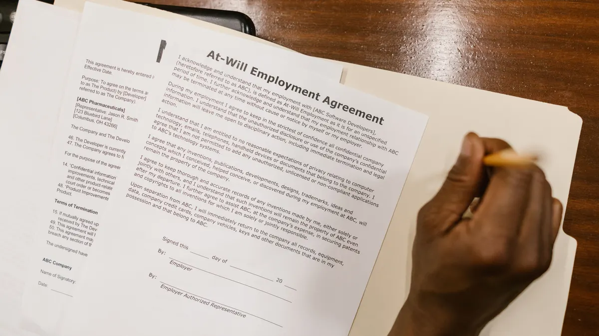 Gros plan d'une main signant un contrat de travail sur une feuille blanche, sur un bureau en bois.