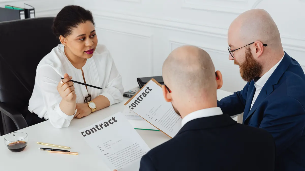 Trois professionnels des affaires discutent d'un contrat dans un bureau moderne ; une femme assise explique les contrats.